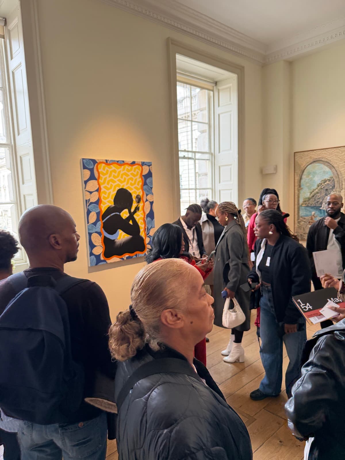 Modern Black Art Club members gathered in a gallery room at 1-54, Somerset House, looking at a painting of a figure in silhouette against a yellow patterned ground.